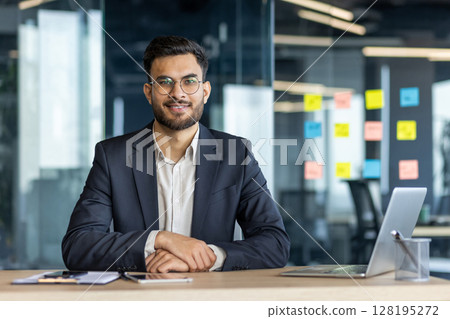A smiling businessman in a modern office setting, seated at a desk, looking directly at the camera. 128195272