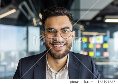 Portrait of successful young businessman at workplace inside office. Man smiling close up and looking at camera, satisfied financier investor. Portrait of successful young businessman at workplace inside office. Man smiling close up and looking at camera, satisfied financier investor. 128195287