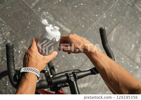 Close up of hands of cyclist holding water bottle, standing with his bike in park on sunny day. Man takes a break from his bike ride. Close up. 128195356