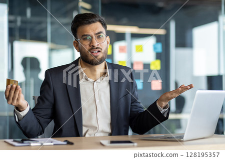A confused businessman looks at a credit card in his hand while working on a laptop in an office environment. The man is expressing financial uncertainty. 128195357