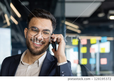 A smiling man with glasses speaks on his phone in an office setting, enjoying his conversation. 128195372