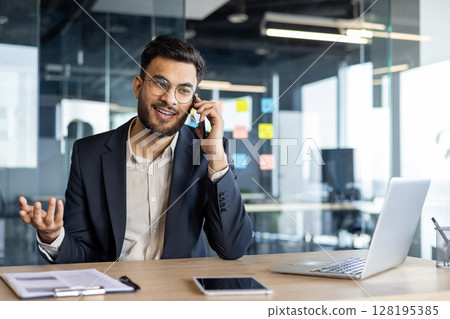 A young businessman talks on the phone while working in a modern office. 128195385