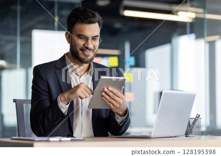 A mature man in formal business attire works confidently using a tablet device. He is seated at his desk in an office with a relaxed yet focused demeanor, surrounded by work tools. A mature man in formal business attire works confidently using a tablet device. He is seated at his desk in an office with a relaxed yet focused demeanor, surrounded by work tools. 128195398