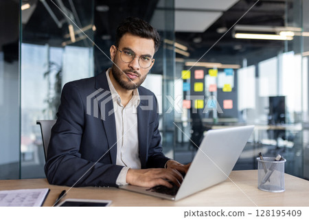 Confident Latin American businessman using a laptop while seated in a contemporary office environment. Focused professional showcasing productivity and determination in a workspace. Confident Latin American businessman using a laptop while seated in a contemporary office environment. Focused professional showcasing productivity and determination in a workspace. 128195409