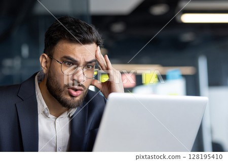 Perplexed businessman in glasses looking at laptop screen in office, showing confusion. Emotion of surprise and stress highlights concept of problem-solving in business environment. Perplexed businessman in glasses looking at laptop screen in office, showing confusion. Emotion of surprise and stress highlights concept of problem-solving in business environment. 128195410
