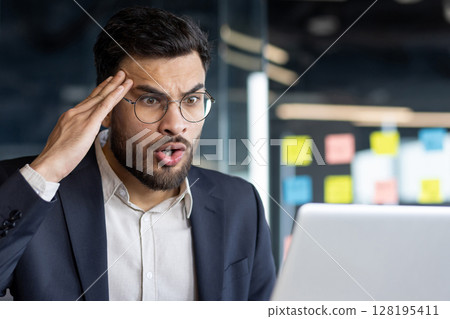 Upset suffering man at workplace inside office. Businessman unhappy and dissatisfied looking at laptop screen, reading online email with bad news. Upset suffering man at workplace inside office. Businessman unhappy and dissatisfied looking at laptop screen, reading online email with bad news. 128195411