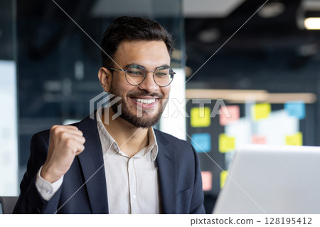 Businessman celebrating success on laptop. Happy professional in office environment expressing triumph. Person excited about achievement, wearing suit and glasses, in work setting. 128195412