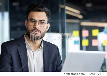 An entrepreneur is working on his laptop in a modern office. He has a determined look and wears a suit jacket over a dress shirt 128195415