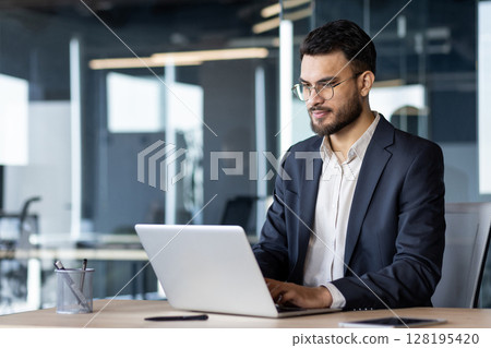 Businessman in modern office using laptop, focused on corporate task. Professional attire and serious expression convey dedication and concentration on work project. Glasses highlight attention. 128195420