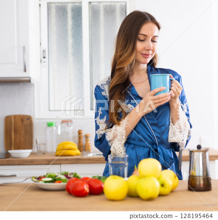 Young woman in blue dressing gown enjoying morning coffee in kitchen Young woman in blue dressing gown enjoying morning coffee in kitchen 128195464