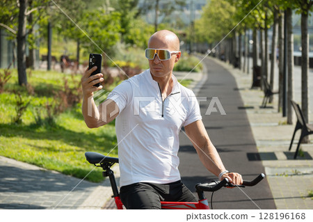 Handsome caucasian adult cyclist taking selfie or talk and video by smart phone in park. Outdoor activity. Handsome caucasian adult cyclist taking selfie or talk and video by smart phone in park. Outdoor activity. 128196168