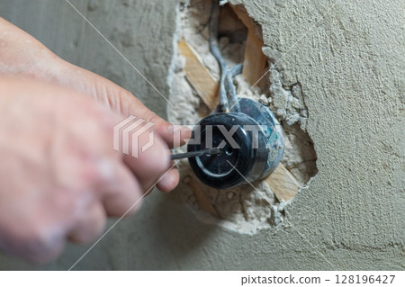 An electrician uses a screwdriver to tighten screws on an electrical outlet during renovation work on a home under construction, ensuring safety and functionality. An electrician installs an outlet. An electrician uses a screwdriver to tighten screws on an electrical outlet during renovation work on a home under construction, ensuring safety and functionality. An electrician installs an outlet. 128196427