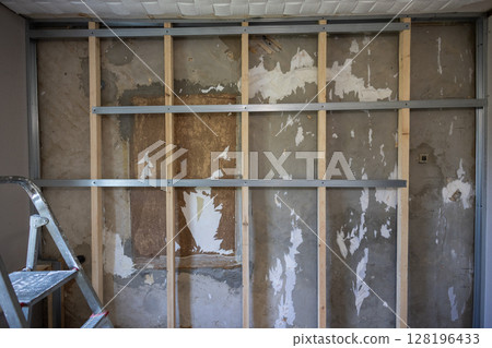 Metal and wood studs are attached to a concrete wall to create a frame for a partition during a home renovation project, with remnants of old wallpaper visible. A frame for attaching drywall sheets. 128196433