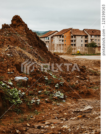 Pile Of Dirt And Debris In A Construction Site Near Residential Buildings: A Muddy Urban Landscape With Scattered Trash, Green Plants, And A Cloudy Sky In The Background 128199163