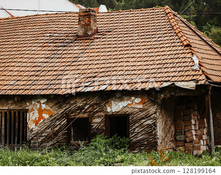 Collapsing Wooden House With A Broken Roof And Crumbling Walls: An Abandoned Rural Structure Overgrown With Greenery And Wildflowers In A Natural Landscape Collapsing Wooden House With A Broken Roof And Crumbling Walls: An Abandoned Rural Structure Overgrown With Greenery And Wildflowers In A Natural Landscape 128199164