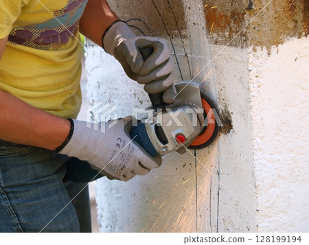 Worker Using an Angle Grinder to Cut Concrete: Sparks Flying as Construction Worker in Gloves and Yellow Shirt Operates Power Tool on Rough Surface 128199194