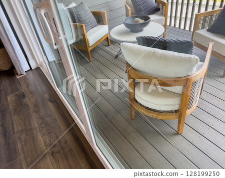 Cushioned patio chairs and a table are visible through a glass sliding door, with a dark composite deck underfoot. The perspective offers an inside-out view of a cozy backyard setup. Cushioned patio chairs and a table are visible through a glass sliding door, with a dark composite deck underfoot. The perspective offers an inside-out view of a cozy backyard setup. 128199250