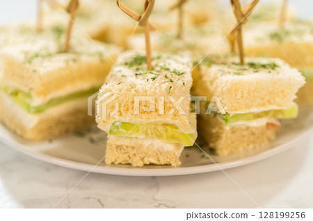 Soft white bread squares layered with a creamy cheese spread and crisp cucumber slices, topped with dried herbs and secured with bamboo cocktail picks. Perfect for elegant tea parties, bridal showers 128199256