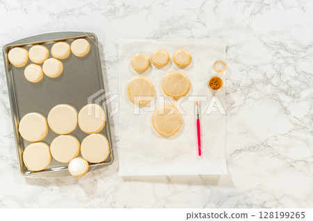 A baking sheet filled with round sugar cookies covered in smooth royal icing, ready for decorating. A brush, edible gold dust, and paper towels sit nearby for the final touches. A baking sheet filled with round sugar cookies covered in smooth royal icing, ready for decorating. A brush, edible gold dust, and paper towels sit nearby for the final touches. 128199265