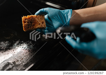 Professional kitchen worker wearing blue gloves spraying detergent on sponge, preparing to clean glass door of modern oven in kitchen with dark interior. Concept of cleanliness during routine chores. Professional kitchen worker wearing blue gloves spraying detergent on sponge, preparing to clean glass door of modern oven in kitchen with dark interior. Concept of cleanliness during routine chores. 128199267