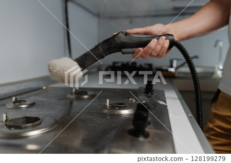 Closeup of housekeeper cleaning dirty gas stove using steam cleaner and cloth, removing grease and burnt food residues, ensuring hygienic kitchen environment. Concept of modern housekeeping appliance. 128199279