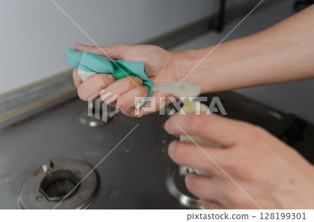 Close-up of housekeeper hands squeezing liquid cleaner from bottle onto sponge, preparing to clean dirty stovetop, emphasizing domestic chores and hygiene. Concept of household chores and hygiene. Close-up of housekeeper hands squeezing liquid cleaner from bottle onto sponge, preparing to clean dirty stovetop, emphasizing domestic chores and hygiene. Concept of household chores and hygiene. 128199301