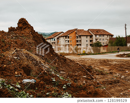 Pile Of Dirt And Debris In A Construction Site Near Residential Buildings: A Muddy Urban Landscape With Scattered Trash, Green Plants, And A Cloudy Sky In The Background 128199329