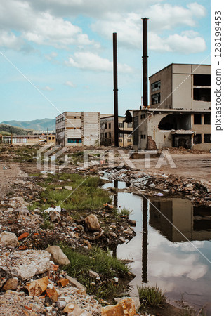 Abandoned Industrial Landscape With Ruins And Reflections: Derelict Factories, Broken Windows, And Overgrown Grass Surrounding Puddles Reflecting Smokestacks And The Sky In A Scene Of Urban Decay Abandoned Industrial Landscape With Ruins And Reflections: Derelict Factories, Broken Windows, And Overgrown Grass Surrounding Puddles Reflecting Smokestacks And The Sky In A Scene Of Urban Decay 128199453