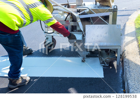 Worker uses specialized machine to paint white line on freshly laid asphalt while wearing safety gear. 128199538