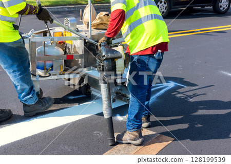 Workers are painting white road lines on an asphalt surface while traffic passes by under clear skies. 128199539