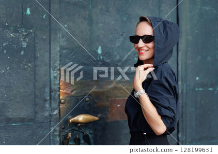 Portrait young adult elegant woman black dress and sunglasses smiles confidently in golden sunlight warm sunset backlit light against historic cathedral building. Female person walking city street Portrait young adult elegant woman black dress and sunglasses smiles confidently in golden sunlight warm sunset backlit light against historic cathedral building. Female person walking city street 128199631