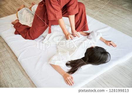 Masseuse making thai yoga massage. Cropped view of woman therapist doing traditional massage treatment for female patient. Client lying on mat, meditating, enjoying hand massage in calm atmosphere. 128199813