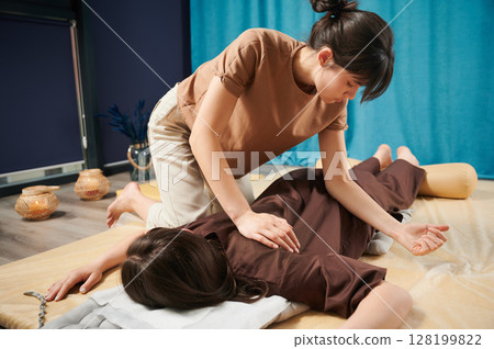 Masseuse making thai yoga massage. Woman therapist doing traditional massage treatment for female patient using elbow. Client lying on mat, meditating, enjoying hand massage in calm atmosphere. 128199822
