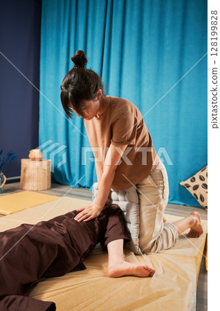 Masseuse making thai yoga massage. Woman therapist doing traditional massage treatment, stretching female patient's body. Client lying on mat, enjoying hand massage in calm atmosphere. 128199828