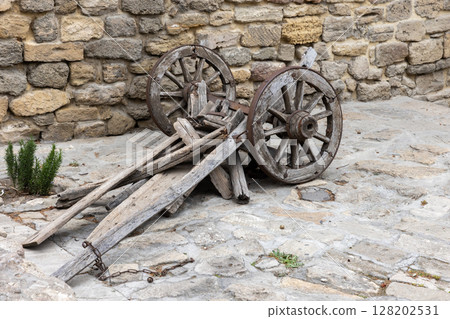 Rustic wooden cart wheels and a wooden chassis lean against a stone wall 128202531