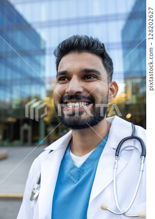 A smiling male doctor with a stethoscope looks away from the camera near a glass building, during daytime. A smiling male doctor with a stethoscope looks away from the camera near a glass building, during daytime. 128202627