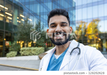 A smiling doctor stands in front of a modern building, wearing a lab coat and stethoscope. He looks directly at the camera. 128202628