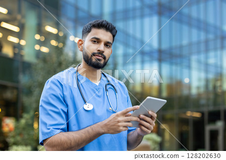 A healthcare professional in scrubs uses a tablet outdoors, in front of a modern building. He wears a stethoscope. 128202630