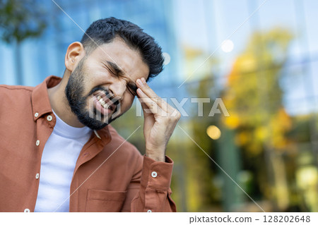 A man holds his head in pain, showcasing distress. Blurred background with building and trees. A man holds his head in pain, showcasing distress. Blurred background with building and trees. 128202648