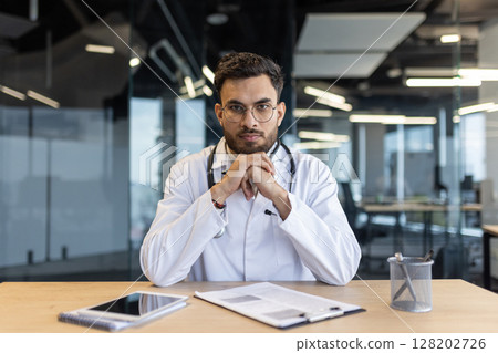Mature male doctor seated at office desk, wearing a stethoscope and white coat, exuding professionalism and focus. The setting depicts a modern medical office Mature male doctor seated at office desk, wearing a stethoscope and white coat, exuding professionalism and focus. The setting depicts a modern medical office 128202726