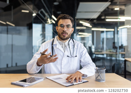 Webcam view, doctor consults patients remotely. Man in white medical coat, uses video call, for online conference meeting. Serious doctor explains course of treatment. Webcam view, doctor consults patients remotely. Man in white medical coat, uses video call, for online conference meeting. Serious doctor explains course of treatment. 128202730