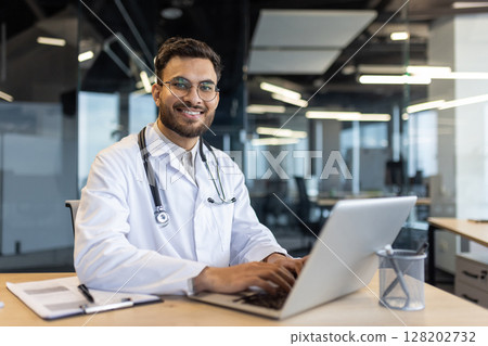 A smiling doctor, wearing a white coat and stethoscope, works on a laptop at a desk in a modern office setting. A smiling doctor, wearing a white coat and stethoscope, works on a laptop at a desk in a modern office setting. 128202732