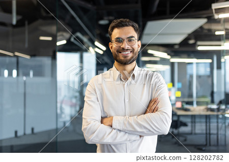 Portrait of successful young businessman man smiling and looking at camera with crossed arms. Office worker satisfied with results of achievement at workplace. 128202782