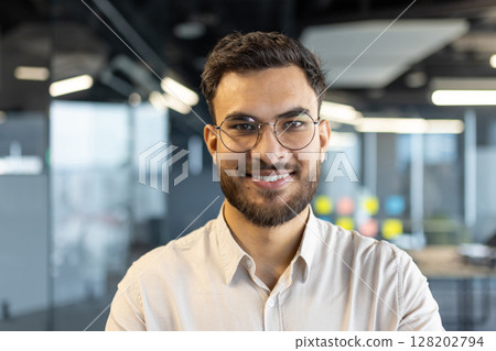 A smiling businessman wearing glasses poses in a modern office setting, showcasing a confident and professional image. A smiling businessman wearing glasses poses in a modern office setting, showcasing a confident and professional image. 128202794