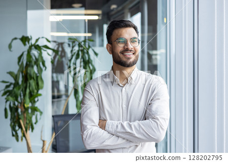 Confident young man in office standing by window, smiling and wearing glasses. Shows positivity, determination, and professionalism in modern business environment. Confident young man in office standing by window, smiling and wearing glasses. Shows positivity, determination, and professionalism in modern business environment. 128202795