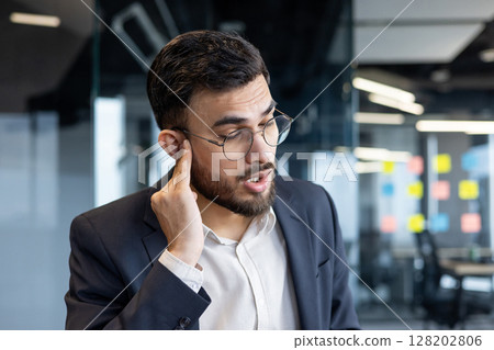 Mature man businessman with glasses experiencing ear pain, possibly due to inflammation or otitis, located in a modern corporate office. The professional attire and modern office background Mature man businessman with glasses experiencing ear pain, possibly due to inflammation or otitis, located in a modern corporate office. The professional attire and modern office background 128202806