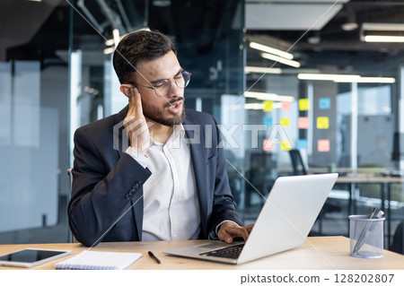 A man in a suit, experiencing ear pain while working on a laptop in an office setting A man in a suit, experiencing ear pain while working on a laptop in an office setting 128202807