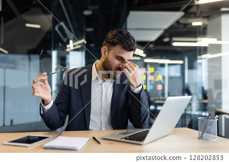 A businessman in a suit, experiencing a headache, takes off his glasses at his desk. He is using a laptop, likely working hard, and is feeling stressed. A businessman in a suit, experiencing a headache, takes off his glasses at his desk. He is using a laptop, likely working hard, and is feeling stressed. 128202853