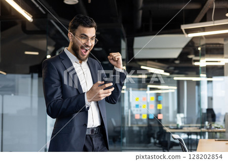 Mature professional businessman expressing joyous emotions after receiving good news on his smartphone. Wearing a formal suit, he conveys an energetic and successful business attitude 128202854