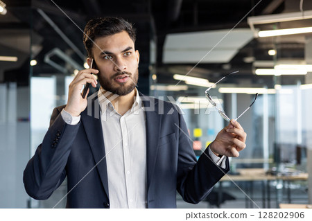 Businessman taking important phone call in office. Holds glasses while conversing, conveys confidence, communication, professionalism. Modern business environment. Tags: businessman, phone, office. Businessman taking important phone call in office. Holds glasses while conversing, conveys confidence, communication, professionalism. Modern business environment. Tags: businessman, phone, office. 128202906